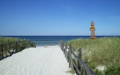 Piękna, piaszczysta lubiatowo plaża z drewnianym płotkiem i wydmami. W oddali widać morze i kilka osób kąpiących się.
