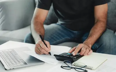 Mężczyzna liczy na kalkulatorze i pisze, być może rozlicza niezgłoszona darowizna od rodziców. Obok laptop, okulary i notatnik.