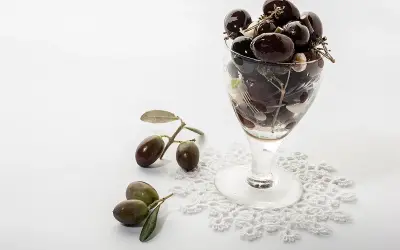 Un bicchiere pieno di olive nere, con rametti e foglie, accanto a olive fresche su un pizzo. Le olive delle Marche, un sapore autentico.