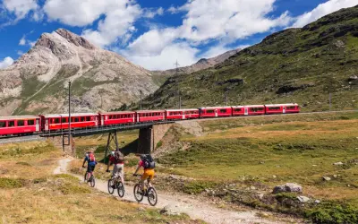 Treno rosso attraversa un ponte su un paesaggio alpino mozzafiato. Tre ciclisti pedalano su un sentiero sottostante, godendosi la vista della ferrovia del Bernina.