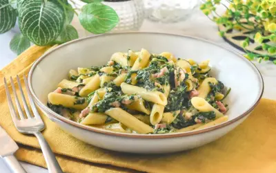 Un plato de pasta penne con espinacas y trocitos de jamón, servido en un cuenco sobre una servilleta amarilla.