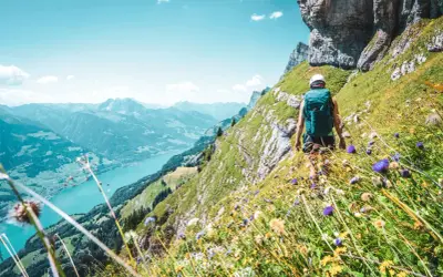 Vandra i Schweiz längs en bergssluttning med en turkos sjö nedanför. Blommor i förgrunden, en vandrare med ryggsäck på väg uppåt.