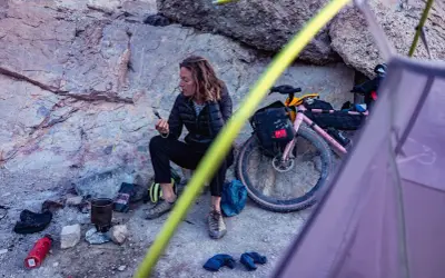 Pause sur une sortie gravel : une cycliste se repose près de son vélo rose, une tasse à la main, au milieu d'un paysage rocheux.