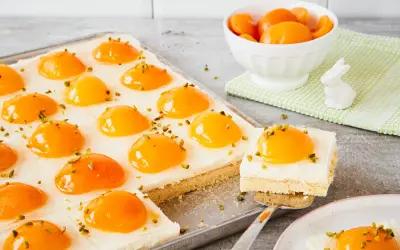 Ciasto z brzoskwiniami, przypominające jajko sadzone, posypane pistacjami. Na blasze i talerzyku.
