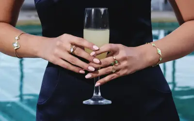 Manos con uñas rosadas y varios anillos dorados, sosteniendo una copa de champán. Brazaletes dorados adornan ambas muñecas.