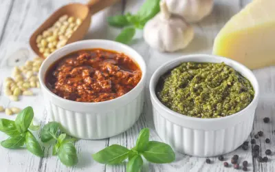 Dwa rodzaje pesto, zielone i czerwone, w białych miseczkach. Obok składniki: orzeszki piniowe, czosnek, bazylia, parmezan. Zastanawiasz się, ile czasu można przechowywać domowe pesto w lodówce?