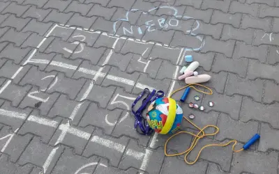 Kreda rysuje podwórkowe zabawy z piłką, skakanką i kredą. Chmurka z napisem "NIEBO" zaprasza do gry.