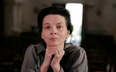 Portret kobiety w wieku dojrzałym, przypominającej Camille Claudel, z dłońmi podpartymi pod brodą, w stonowanym wnętrzu.