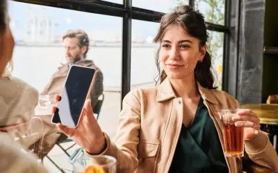 Kobieta trzyma telefon, słuchając wiadomości na poczcie głosowej. W tle mężczyzna i widok na port.