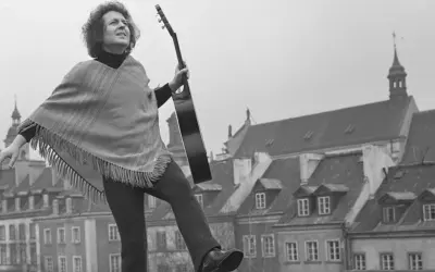 Tadeusz Woźniak, zegarmistrz światła, z gitarą na tle kamienic.