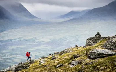En person med röd ryggsäck vandrar uppför en stenig sluttning med vidsträckt, dimmig fjällvy. Abisko vandring i sitt esse.