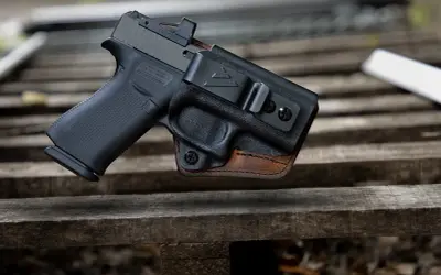 Pistolet w kaburze udowej z czarnego polimeru i brązowej skóry, z widocznym celownikiem optycznym.