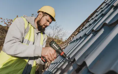 A roofer in a yellow hard hat and hi-vis vest uses a drill for metal roof repair.