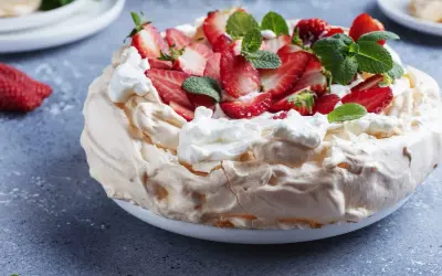 Pyszny tort bezowy z mascarpone i bitą śmietaną, ozdobiony świeżymi truskawkami i miętą.