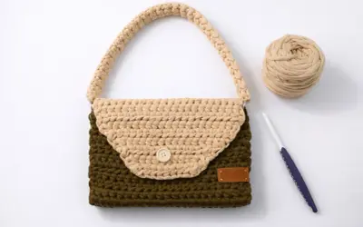 Bolso de trapillo bicolor, verde y beige, con asa y botón de madera. A su lado, ovillo de lana y ganchillo.