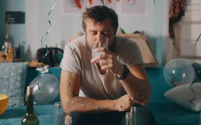 Zmęczony mężczyzna z tatuażami pije wodę po imprezie, symbolizującą jego odstawienie alkoholu. W tle resztki zabawy.