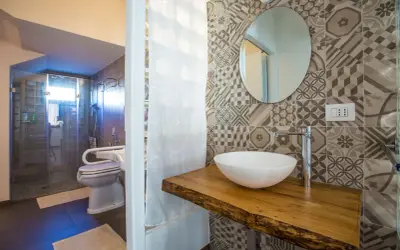 Bagno piccolo con antibagno, doccia in vetro, WC e lavabo su mensola in legno grezzo. Piastrelle geometriche e specchio rotondo.