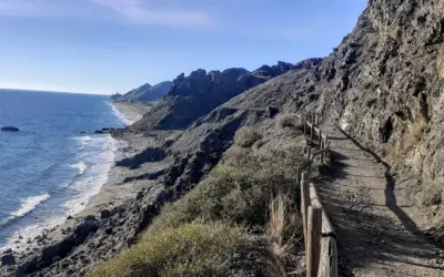 Sendero costero con vistas al mar Mediterráneo y montañas. Un camino de tierra con barandilla de madera serpentea por la ladera, ideal para un paseo marítimo en Mojácar Playa.