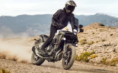 Motocyklista w kasku i kurtce pędzi na motocyklu Honda CB500X po piaszczystej drodze, wzbijając tumany kurzu.