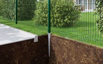 Montaż słupków ogrodzeniowych instrukcja: zielony słupek z metalową stopą wbity w ziemię, obok betonowy chodnik i siatka ogrodzeniowa.