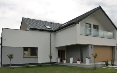 Nowoczesny dom z dwuspadowym dachem i balkonem. Elewacja w odcieniach bieli i szarości, z drewnianą bramą garażową. Świetny pomysł na elewację domu.