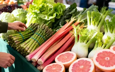 Un étal de marché débordant de fruits et légumes de saison : asperges, rhubarbe, fenouils, laitues et pamplemousses coupés.