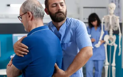 Fizjoterapeuta pomaga starszemu mężczyźnie z bólem pleców. W tle widać szkielet i drugą pielęgniarkę.