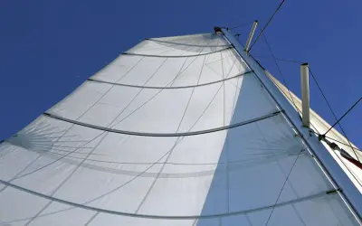 Vela bianca randa full batten spiegata al vento, con stecche orizzontali e cavi che salgono verso il cielo azzurro.