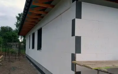 Dom w trakcie budowy, widoczna biała izolacja ścian zewnętrznych z szarymi narożnikami. Dach z drewnianymi krokwiami.