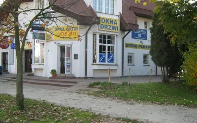 Budynek z żółtymi banerami "OKNA DRZWI" i "DRZWI PARAPETY", z drzewem po lewej.