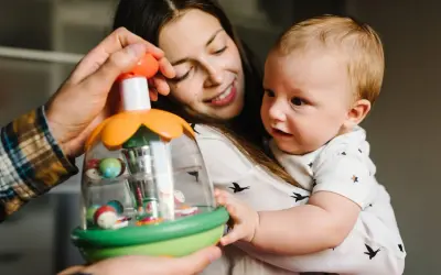 Mama i tata pokazują dziecku zabawkę. Maluch, choć nie jest jeszcze dwulatkiem, z zaciekawieniem kręci głową, obserwując kolorowe kulki.