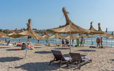 Słoneczna plaża z parasolami z trawy, leżakami i ludźmi cieszącymi się wakacjami. To idealne miejsce na **carewo atrakcje**.