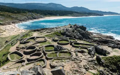 Ruinas circulares de un castro gallego junto a una playa de aguas turquesas. Paisajes de Galicia que evocan historia y naturaleza.