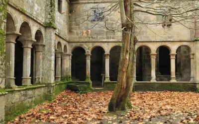 Claustro de piedra cubierto de musgo, con arcos y columnas, rodeado de hojas secas. Un árbol solitario se alza en primer plano, evocando la atmósfera de los monasterios de Galicia.