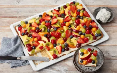 Bunter Blechkuchen mit Obst, belegt mit Erdbeeren, Kiwi, Trauben, Pfirsichen und Beeren. Ein Stück ist bereits auf einem Teller angerichtet.