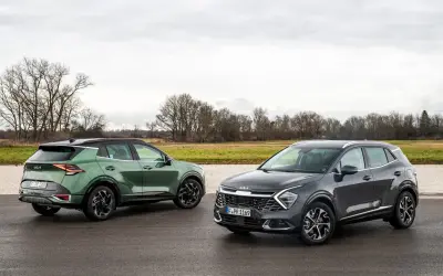 Dwa nowoczesne crossovery Kia Sportage, zielony i grafitowy, stoją na mokrej drodze na tle jesiennego krajobrazu.