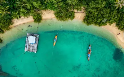 Drohne über Thailand: Ein Blick von oben auf eine schwimmende Plattform und zwei Boote im türkisfarbenen Meer, umgeben von üppiger Vegetation und Sandstrand.