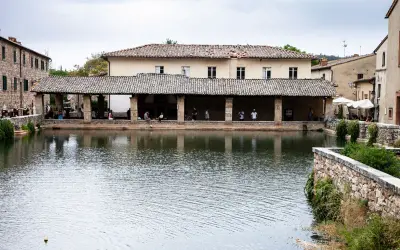 La piazza delle sorgenti termali di Bagni Vignone, con persone che si rilassano sotto un portico e il riflesso dell'acqua.