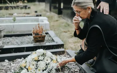 Kobieta w żałobie klęczy przy grobie, ocierając łzy. Bukiet kwiatów leży na ziemi. To smutne ostatnie pożegnanie.