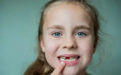 Uśmiechnięta dziewczynka z niebieskimi oczami pokazuje lukę po wypadniętym mleczaku. To znak, że czas na nowe zęby!