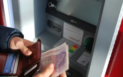 Person hält Geld und Portemonnaie vor Geldautomat. Hier kann man bei der Bank Geld wechseln.