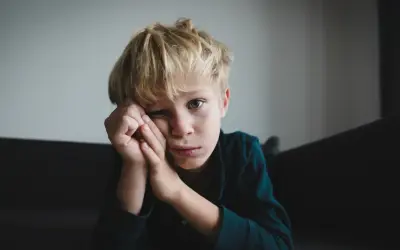 Chłopiec z blond włosami, zamyślony, opiera głowę na dłoniach. Jego spojrzenie sugeruje trudności, być może związane z zespołem Aspergera.