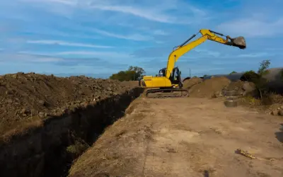 Excavadora amarilla trabajando en la creación de varios tipos de zanjas, con montones de tierra y tuberías grandes en el fondo.
