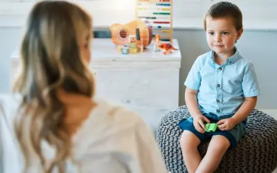 Chłopiec w niebieskiej koszuli siedzi na pufie, rozmawiając z psychoterapeutką.