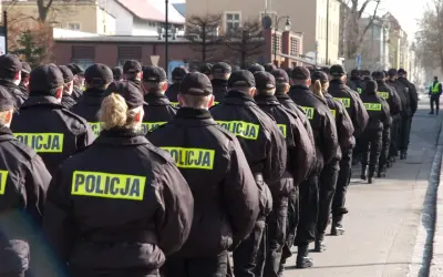 Policjanci w czarnych kurtkach z napisem "POLICJA" stoją w szeregu, gotowi do akcji. Widać, jak pracuje policja zmiany, przygotowując się do służby.