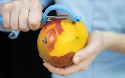 Hände schälen eine reife Mango mit einem blauen Sparschäler. Das Fruchtfleisch leuchtet gelb, während die Schale rot und gelb ist.