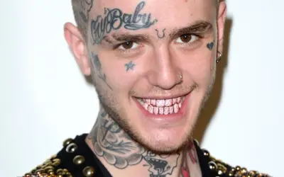 Uśmiechnięty Lil Peep z tatuażami na twarzy i szyi, błyszczącymi różowymi zębami. Pytanie czy lil peep żyje, pozostaje w sercach fanów.
