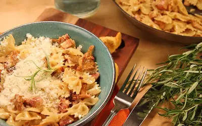 En skål med krämig kantarellpasta med strimlad ost och en kvist rosmarin. En panna med samma pasta syns i bakgrunden.