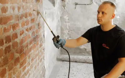 Mann sprüht mit Gerät gegen Feuchtigkeit im Keller. Ziegelwand wird behandelt.