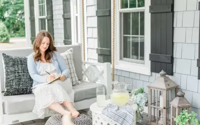 A woman embodies the coastal grandma aesthetic, writing in a notebook on a porch swing, with lemonade and hydrangeas nearby.